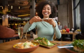 Imagem de uma mulher sorridente sentada à mesa de um restaurante tirando uma foto de sua comida com o celular, cercada por pratos coloridos e uma bebida em um ambiente moderno e aconchegante, para ilustrar matéria sobre os melhores restaurantes em Nova Iguaçu
