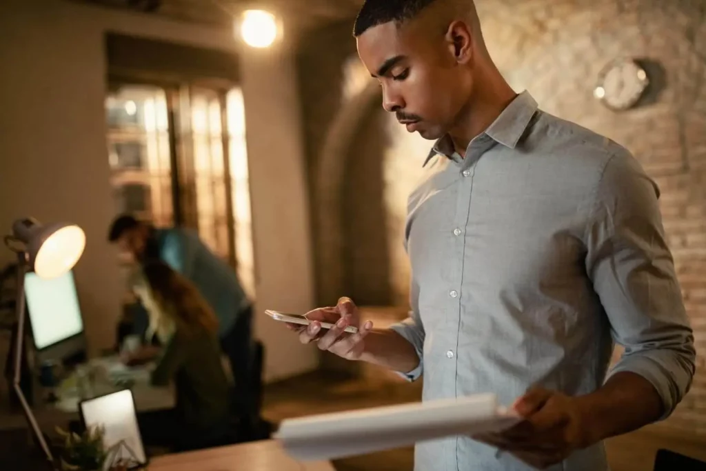 Imagem de umhomem de camisa cinza-clara em um escritório, olhando para o telefone e segurando documentos, para ilustrar matéria sobre o que o subsíndico não pode fazer. Ao fundo, dois colegas de trabalho trabalham em uma mesa com computadores e iluminação quente.