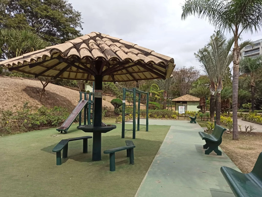 Imagem de um playground ao ar livre com bancos, escorregador e equipamentos de ginástica sob um grande gazebo no Parque Cássia Eller, em Belo Horizonte, cercado por árvores, plantas e vegetação em um dia nublado. Um caminho e uma placa são visíveis ao fundo.