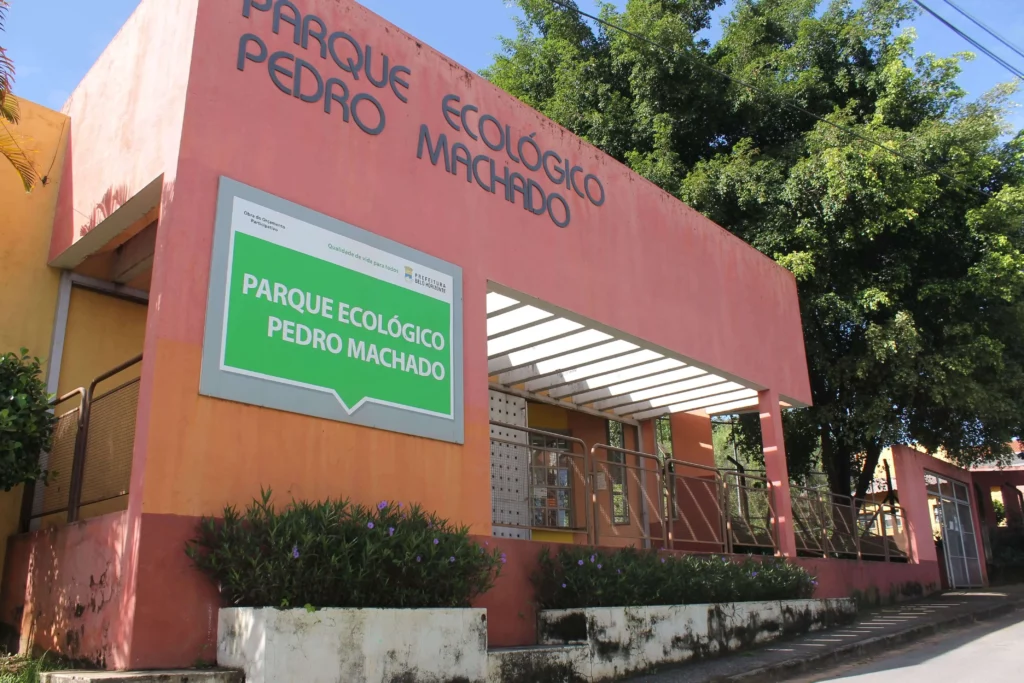 Imagem da entrada do Parque Ecológico Pedro Machado, com uma grande placa e um prédio rosa-alaranjado cercado por plantas e árvores, em um dia ensolarado.