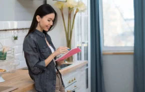 Imagem de uma mulher em pé em uma cozinha sorrindo enquanto olha e escreve em um caderno rosa para ilustrar matéria sobre o que significa subsíndico. Ela está vestindo uma camisa jeans e tem longos cabelos escuros. A cozinha tem decoração moderna, flores e luz natural de uma janela.