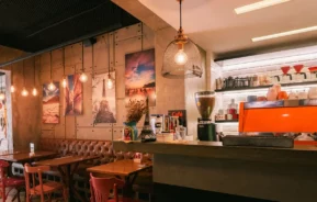 Um café aconchegante com iluminação quente, mesas de madeira, cadeiras vermelhas e marrons, arte na parede e um balcão com uma máquina de café expresso laranja brilhante e prateleiras com xícaras de café e equipamentos.