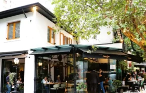 Uma cafeteria moderna com grandes janelas de vidro e um toldo verde fica em uma rua arborizada. As pessoas estão jantando dentro e fora, desfrutando da atmosfera animada sob a sombra das árvores frondosas.