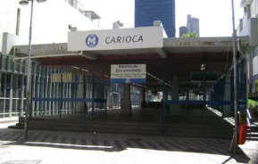 Entrada da estação de metrô Carioca no Rio de Janeiro, Brasil, mostrando uma placa com o nome da estação, painéis de vidro e prédios altos ao fundo em um dia ensolarado.