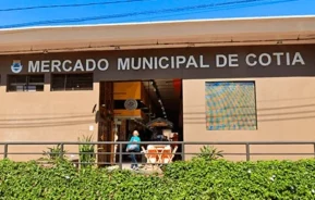 Vista externa do Mercado Municipal de Cotia, um prédio bronzeado com uma grande placa acima da entrada, sob um céu azul claro. É possível ver pessoas entrando e saindo do mercado.