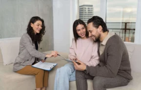 Imagem de uma mulher com uma prancheta falando e gesticulando para um casal sorridente que está sentado em um sofá para ilustrar matéria sobre aluguel de casa. O casal olha para um tablet juntos, parecendo engajados e felizes em um ambiente de escritório moderno e iluminado.