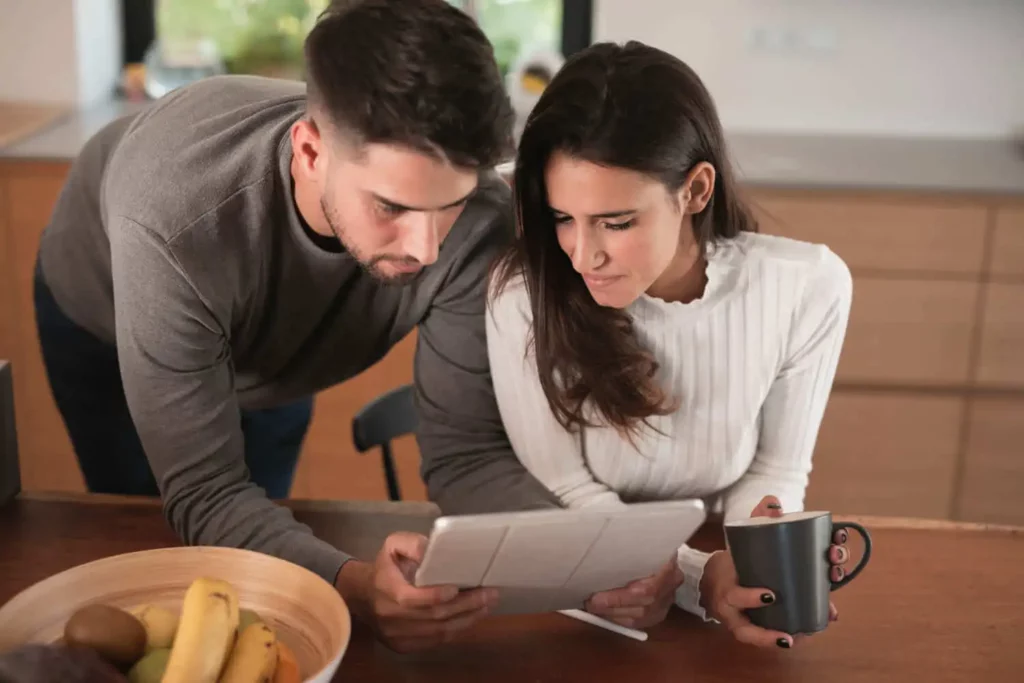 Imagem de um homem e uma mulher sentados em um balcão de cozinha olhando juntos para um tablet para ilustrar matéria sobre aluguel de casa na praia. A mulher segura uma caneca de café e há uma tigela de frutas sobre o balcão em primeiro plano. Eles parecem concentrados e engajados.