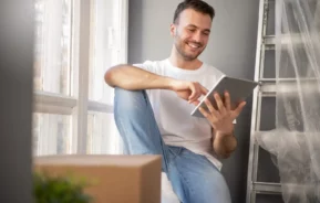 Imagem de um homem sorridente, vestindo camiseta branca e calça jeans, está sentado perto de uma janela, usando um tablet. Uma caixa de papelão e uma escada coberta com plástico estão próximas.