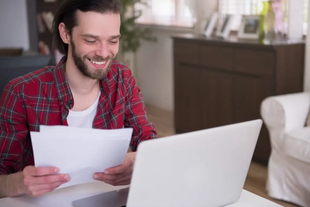Imagem de um homem sorridente com uma camisa xadrez vermelha sentado à mesa, segurando papéis e olhando para a tela de um laptop em uma sala iluminada e aconchegante com prateleiras e um aparador ao fundo.