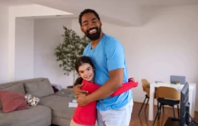 Imagem de um homem sorridente com uma camisa azul-clara abraçando uma criança com uma blusa vermelha em uma sala de estar moderna com um sofá, uma mesa e cadeiras ao fundo.