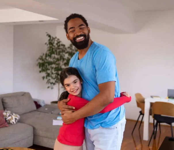 Imagem de um homem sorridente com uma camisa azul-clara abraçando uma criança com uma blusa vermelha em uma sala de estar moderna com um sofá, uma mesa e cadeiras ao fundo.