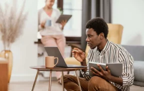 Imagem de um homem sentado em uma pequena mesa, trabalhando em um laptop e segurando um tablet, parecendo concentrado. Uma mulher ao fundo está ao lado de uma janela, segurando uma xícara e um tablet. O cenário parece ser uma sala de estar aconchegante.