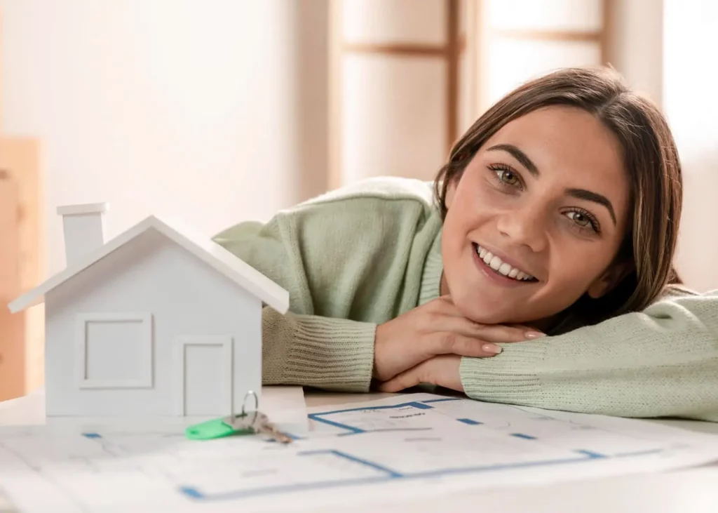 Imagem de uma mulher sorridente apoiada em uma mesa com os braços cruzados, ao lado de um modelo de casa pequena, chaves de casa e plantas de construção.