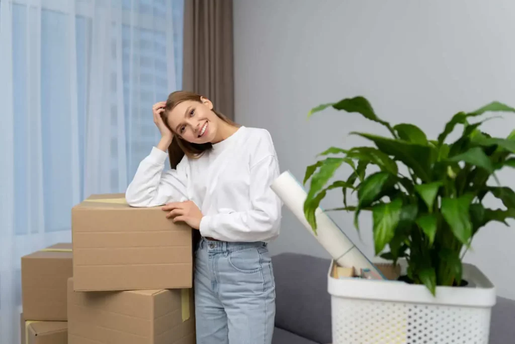 Imagem de uma mulher sorridente, vestindo um suéter branco e calça jeans, inclina a cabeça sobre caixas de papelão empilhadas, com um grande vaso de planta verde e materiais de mudança ao seu lado em uma sala iluminada.