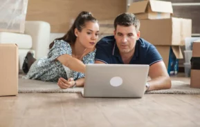 Imagem de um casal deitado no chão, cercado por caixas de mudança, olhando juntos para um laptop para ilustrar matéria sobre inadimplência no consórcio.