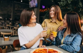 Imagem de três mulheres jovens sentadas ao redor de uma mesa de um restaurante, sorrindo e bebendo copos de suco de laranja, com uma pizza à sua frente, para ilustrar matéria sobre o Mercado da Boca. O ambiente é casual e aconchegante, com iluminação quente e atmosfera descontraída.