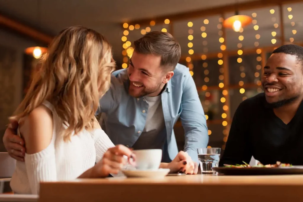Imagem de três amigos desfrutando de uma refeição juntos em um restaurante. Uma pessoa está de pé e sorrindo com a mão no ombro do amigo sentado, enquanto os outros dois estão sentados, conversando e segurando bebidas. Luzes quentes pendem ao fundo.