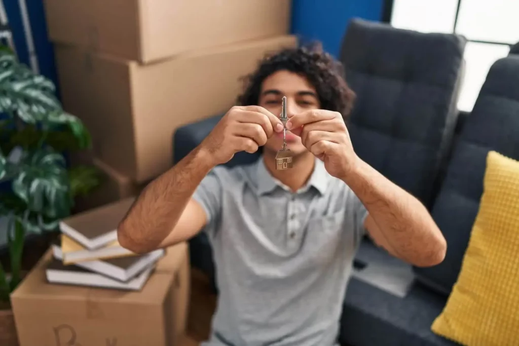 Imagem de um homem na sala de um imóvel segurando as chaves de casa cercadopor caixas de mudança, livros e um sofá com uma almofada amarela.