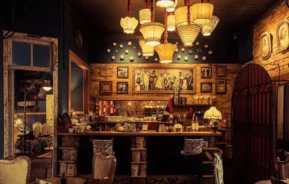 Uma cafeteria vintage aconchegante e pouco iluminada com cadeiras ornamentadas, um bar de madeira, iluminação quente de lâmpadas ecléticas, arte emoldurada em paredes de tijolos e detalhes decorativos que criam uma atmosfera convidativa e nostálgica.
