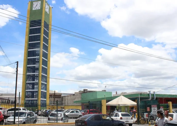 Uma torre de relógio alta, verde e amarela, fica ao lado de um estacionamento cheio de carros sob um céu parcialmente nublado. Pessoas caminham perto de uma cerca verde e de um prédio amarelo ao fundo.