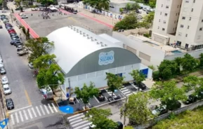 Vista aérea de um grande ginásio denominado "Suzano", cercado por árvores, carros estacionados e edifícios residenciais altos, com um pátio de escola e pessoas visíveis ao fundo.