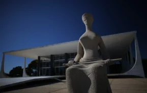 Uma estátua de pedra de uma mulher sentada, com os olhos vendados e segurando uma espada, está em frente a um edifício modernista com grandes colunas e paredes de vidro sob um céu azul claro.