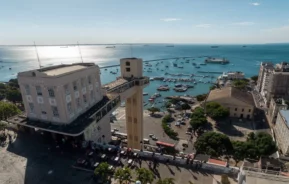 Vista aérea do Elevador Lacerda em Salvador, Brasil, com vista para o porto repleto de barcos, edifícios históricos e oceano azul sob um céu ensolarado.