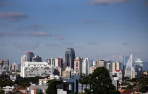 O horizonte de uma cidade com vários prédios modernos sob um céu azul com nuvens dispersas e árvores visíveis em primeiro plano.