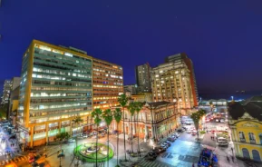 Uma paisagem urbana à noite mostra prédios bem iluminados ao redor de uma pequena fonte com luzes verdes. Os carros estão estacionados ao longo das ruas e as palmeiras se alinham nas calçadas sob um céu azul escuro.