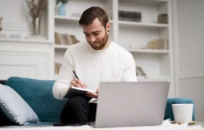 Imagem de um homem de barba =sentado em um sofá azul, sorrindo enquanto escreve em um caderno. Um laptop, uma xícara e alguns biscoitos estão sobre a mesa à sua frente. Ao fundo, há prateleiras com livros.