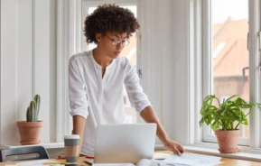 Imagem de uma mulher usando óculos e camisa branca está em pé em frente a uma mesa, olhando documentos. Um laptop, uma xícara de café e um livro aberto estão sobre a mesa. Há dois vasos de plantas e grandes janelas ao fundo.