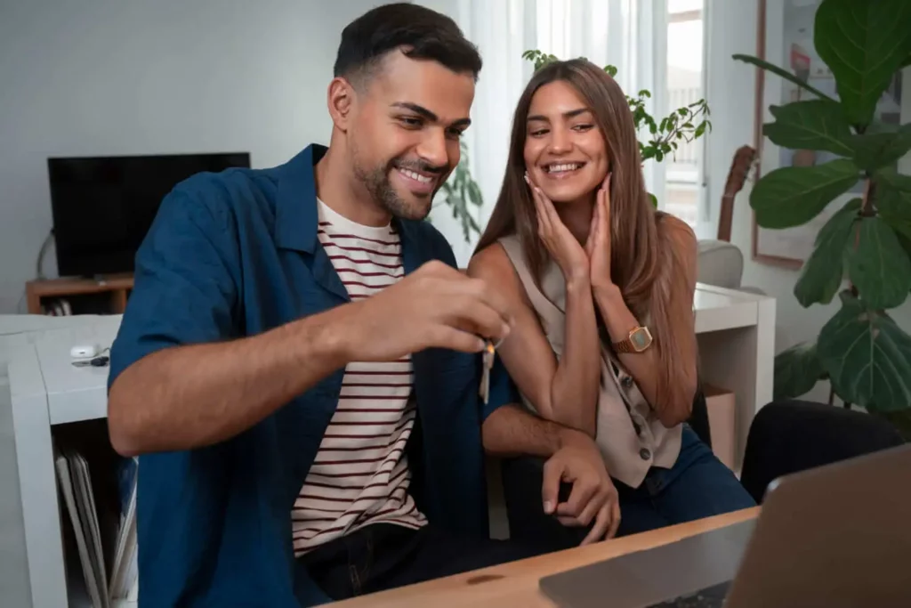 Imagem de um casal sorridente sentado em uma mesa, segurando uma chave na frente de um laptop, parecendo feliz e animado. Eles parecem estar em um ambiente doméstico moderno e iluminado.