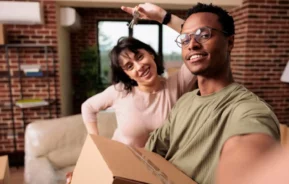 Imagem de um casal sorridente tirando uma selfie enquanto segura caixas de mudança e chaves, em uma aconchegante sala de estar com paredes de tijolos, comemorando a mudança para uma nova casa para ilustrar matéria sobre usar FGTS no consórcio