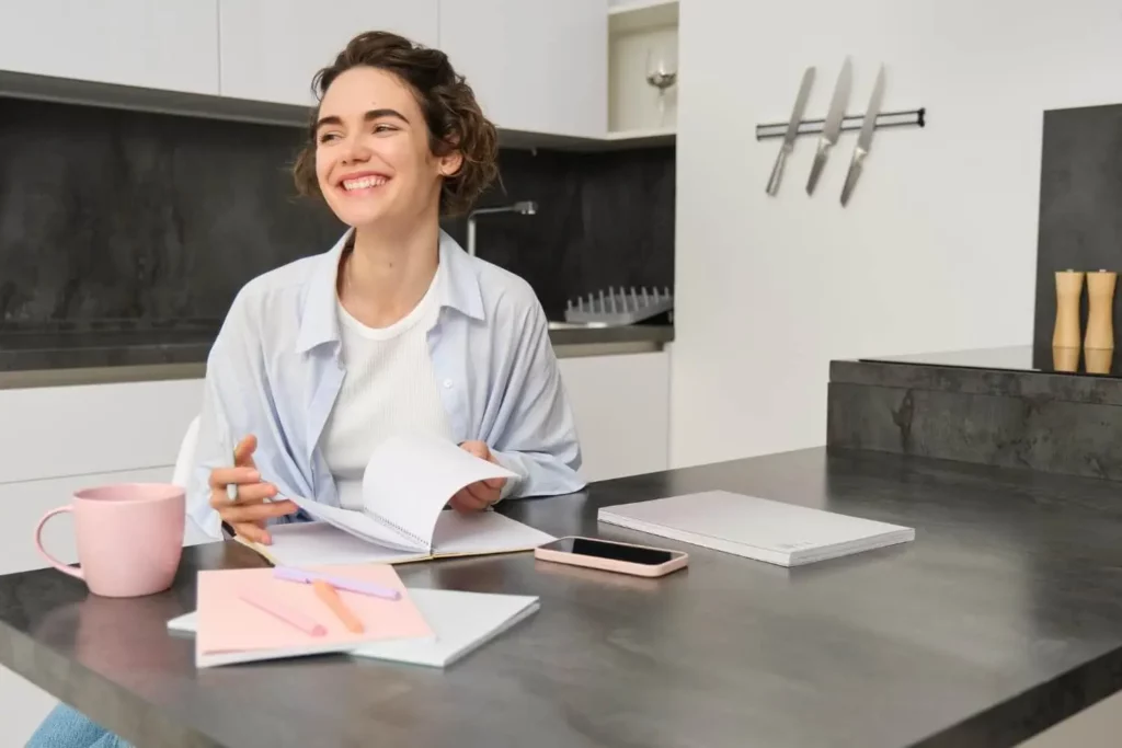 Imagem de uma mulher sorridente sentada em um balcão de cozinha com cadernos, um smartphone e uma caneca rosa. Ela está folheando papéis, parecendo relaxada e feliz, com utensílios de cozinha visíveis no fundo moderno.