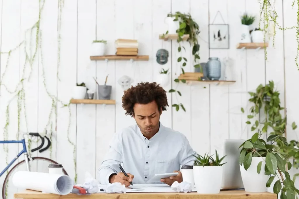 Imagem de um homem de cabelos cacheados sentado em uma escrivaninha cercada de plantas, escrevendo em papéis e olhando para um tablet. A sala tem paredes brancas com prateleiras com livros, plantas e decorações. Uma bicicleta azul está parcialmente visível à esquerda.