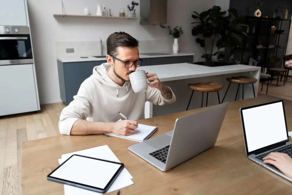 Imagem de um homem de óculos sentado à mesa em uma cozinha moderna, bebendo de uma caneca enquanto escreve em um caderno. Ele está trabalhando em um laptop, com outro laptop e um tablet sobre a mesa