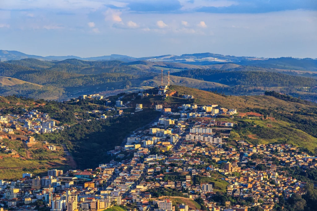 Imagem da vista aérea de Poços de Caldas mostra vários edifícios espalhados por um terreno montanhoso, cercado por colinas verdes e arborizadas, sob um céu parcialmente nublado