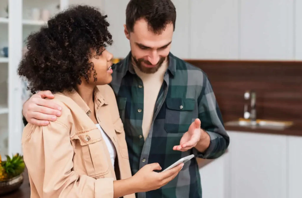 Imagem de uma mulher e um homem juntos em uma cozinha, sorrindo e olhando para um smartphone que a mulher está segurando. O homem gesticula com uma das mãos, e eles parecem estar tendo uma conversa amigável.