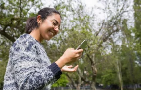 Imagem de uma jovem mulher usando óculos e suéter enquanto usa seu smartphone ao ar livre para ilustrar matéria sobre a Praça da Árvore, em São Paulo. Ela está em um parque com árvores verdes e um céu nublado ao fundo.