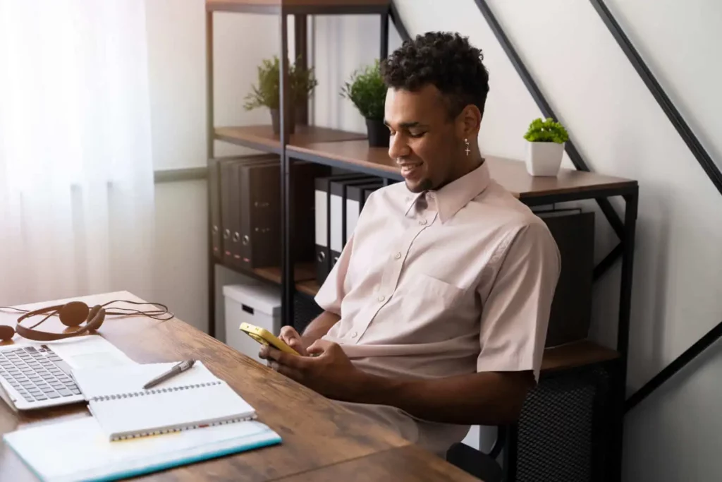 Imagem de um homem sentado em uma mesa com um laptop, um notebook, fones de ouvido e uma caneta, sorrindo enquanto olha para o telefone. Prateleiras com plantas e pastas estão ao fundo, e a luz do sol entra por uma janela.