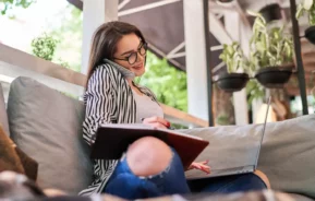 Imagem de uma mulher de óculos sentada em um sofá, falando ao telefone, segurando um caderno e usando um laptop, fazendo várias tarefas em um espaço interno moderno e bem iluminado, com plantas ao fundo.