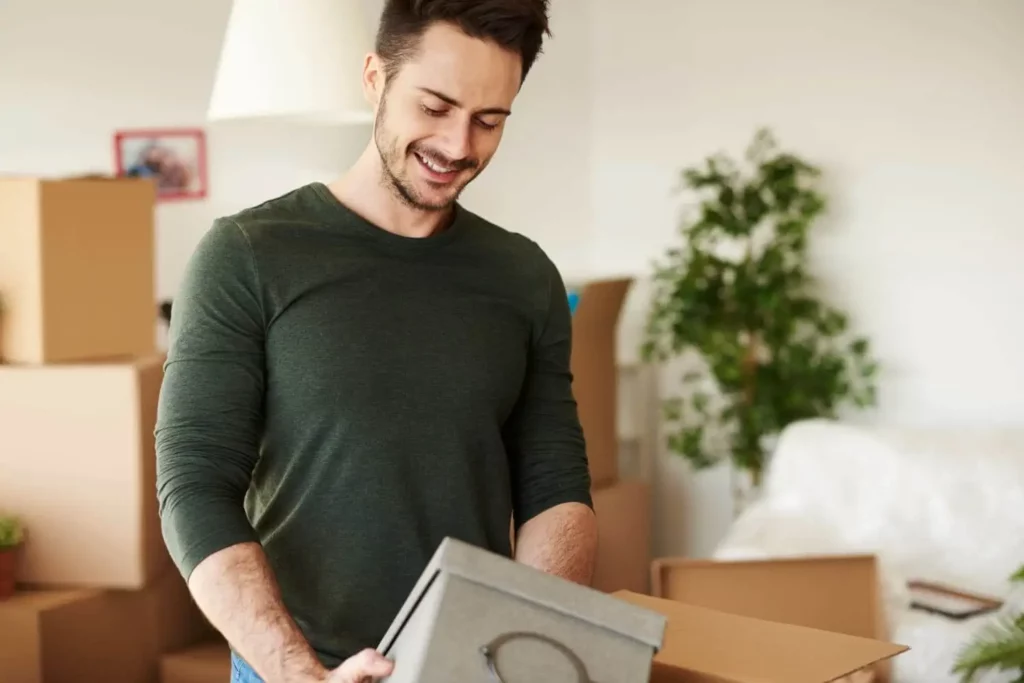 Imagem de um homem sorridente, vestindo uma camisa verde de mangas compridas, segurando uma caixa cinza em uma sala cheia de caixas de papelão e um vaso de plantas ao fundo.