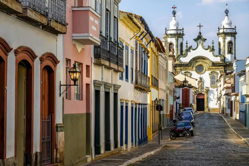 Imagem da paisagem urbana de São João del-Rei mostra casas em estilo colonial, rua de paralelepípedo e igreja ao fundo 