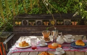 Uma mesa aconchegante ao ar livre preparada para o café da manhã com croissants, xícaras de café, uma prensa francesa e suco. A mesa é coberta com um pano colorido e cercada por vegetação e bancos de madeira decorados com azulejos de borboletas.