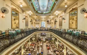 Um restaurante grandioso, de dois andares, com grades ornamentadas, decoração elegante, tetos altos e uma grande claraboia de vitral. Ambos os andares têm mesas de jantar, e o térreo está repleto de pessoas desfrutando de suas refeições.