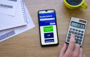 Foto que ilustra matéria sobre como declarar doação de imóvel no Imposto de Renda 2026 mostra uma mão pressiona uma calculadora ao lado de um smartphone que exibe o site da Receita Federal, com papéis, um caderno e uma xícara de café amarela sobre uma mesa de madeira.