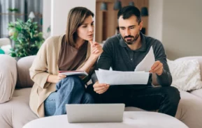 Uma mulher e um homem sentados em um sofá, olhando para papéis e um laptop na mesa à sua frente. A mulher segura um caderno e uma caneta, parecendo pensativa, enquanto o homem examina os documentos.