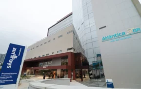 Edifício hospitalar moderno com paredes de vidro e bege. Placas em português mostram Atlântica DOr e Pronto Socorro. As pessoas estão entrando pela entrada principal; a cena se passa durante o dia.