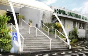 Vista externa do Hospital e Maternidade São Lucas, mostrando uma escada com corrimão, uma rampa para acesso de cadeira de rodas e uma calçada com padrão na frente. O edifício é cinza com sinalização verde e plantas ao redor.