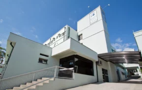 Foto do Hospital Santa Lucinda, em Sorocaba, um edifício moderno, de cor clara, com grandes janelas e uma rampa que leva à entrada, com uma torre com uma placa sob um céu azul claro.